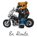 Ursinho estilizado em uma moto, usando jaqueta de couro e óculos escuros, simbolizando liberdade e aventura. Texto "En Route" abaixo da imagem.